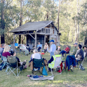 Group sitting outside in a circle in camp chairs with an old wooden building in the background.