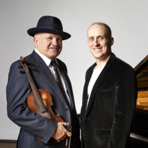 Two musicians, the man on the left holds a violin, on the right is the edge of a piano.