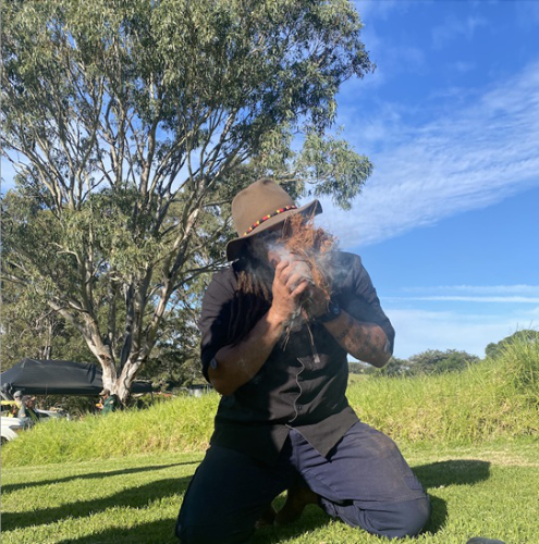 Man kneeling in an open area blowing into tinder in his hands to get the fire started.