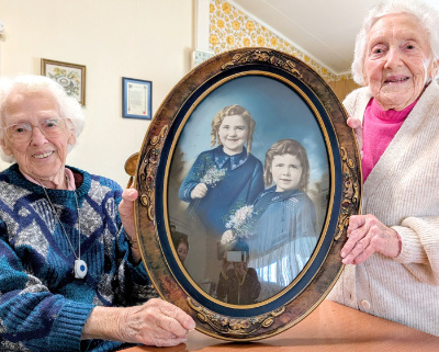 Two older women holding a large oval photo of themselves as young girls.