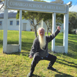 Man in a tai chi pose in front of a sign that says Cobargo School of Arts Hall