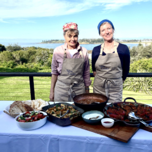 Two women in full cover aprons in front of a table laden with food.