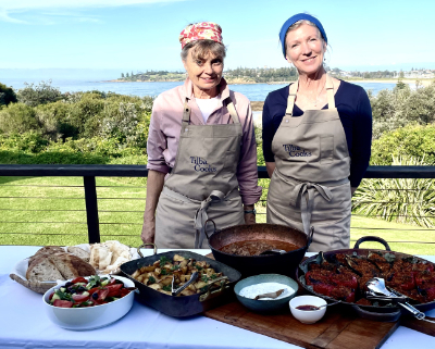 Two women in full cover aprons in front of a table laden with food.