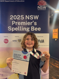 Girl holding her medal and certificate, standing in front of a sign saying '2025 NSW Premier's Spelling Bee'