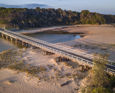 View from the air of a one-lane bridge crossing a sandy estuary.