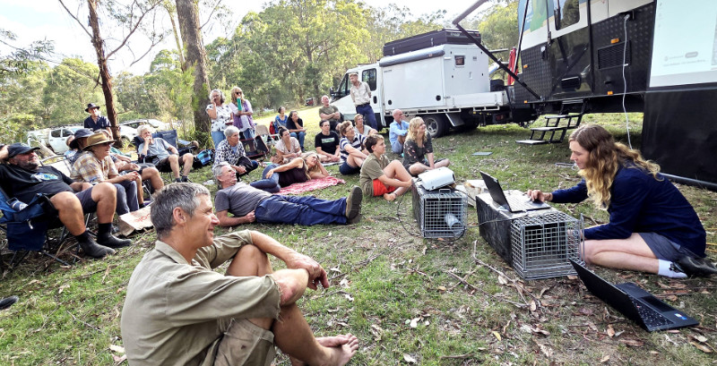 A group seated on the ground in front of a woman giving a presentation.