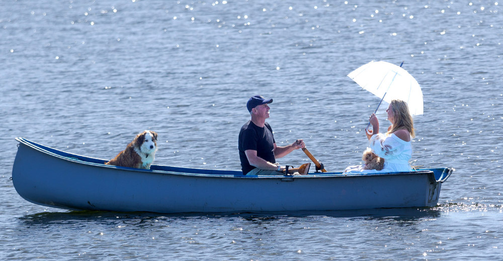 Boat with passengers: dog, man rowing and woman holding a parasol.