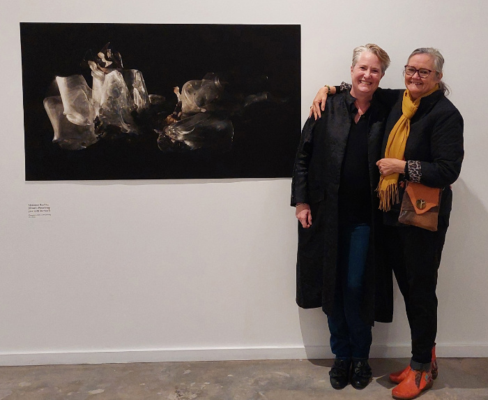 Two women standing beside an artwork on the wall.
