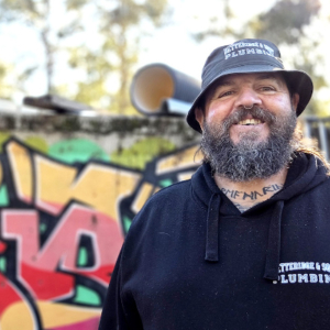 A man with a beard wearing a hat standing in front of a wall with colourful graffiti